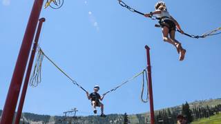 Children bounce high in harnesses at Schweitzer's bungee trampoline, enjoying flips and jumps under a clear summer sky