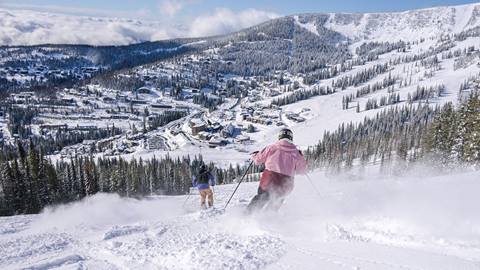 two skiers skiing towards schweitzer village on a sunny powder day