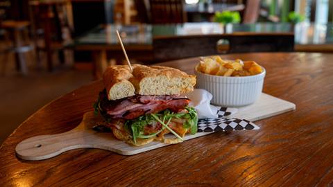 Image of a sandwich with meat, arugula, cheese, and veggies on a board with a bowl of chips at Gourmandie