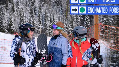 three girls carrying snowboards stand under a trail sign, discussing their kids group snowboard lessons at schweitzer mountain resort