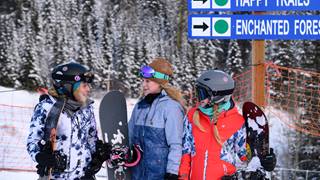 three girls carrying snowboards stand under a trail sign, discussing their kids group snowboard lessons at schweitzer mountain resort