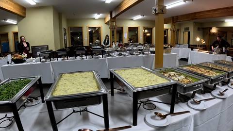 A wide variety of hot dishes are displayed in silver chafing servers on white linen-covered buffet tables for a catered event at Schweitzer.