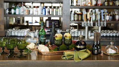 A decorative display of green glassware, floral arrangements, and premium liquor bottles sits on a wooden bar top at Schweitzer.