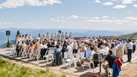A large crowd of guests sits in white chairs during an outdoor wedding ceremony at a mountain summit venue at Schweitzer.