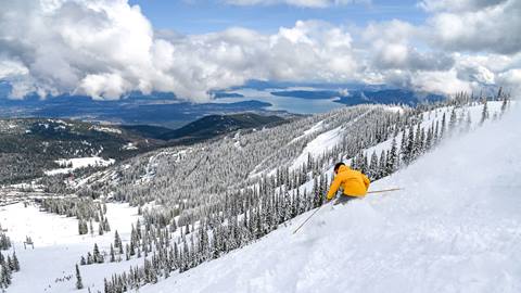 person in yellow coat skiing through powder with a view of lake pend oreille in the background