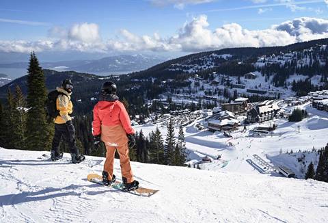 Two snowboarders stand on a snowy ridge at Schweitzer looking out over the mountain village and Lake Pend Oreille.