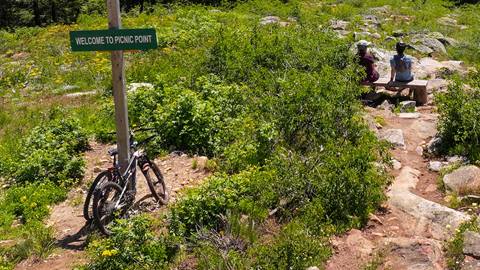 people sitting on a bench with their xc mountain bikes sitting up against a sign that says Picnic point