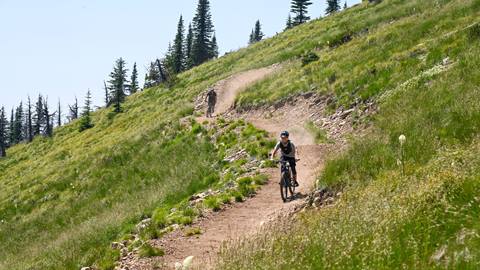 Two mountain bikers descend a high alpine singletrack trail through open grassy slopes with sweeping views