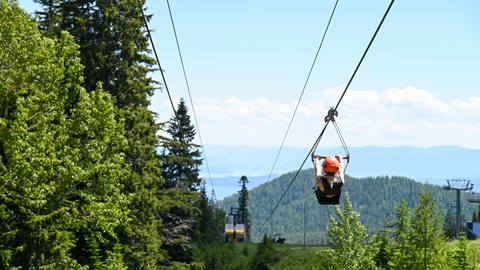 child wearing a helmet riding a zipline through the forest at Schweitzer with scenic views of lake pend oreille in the background